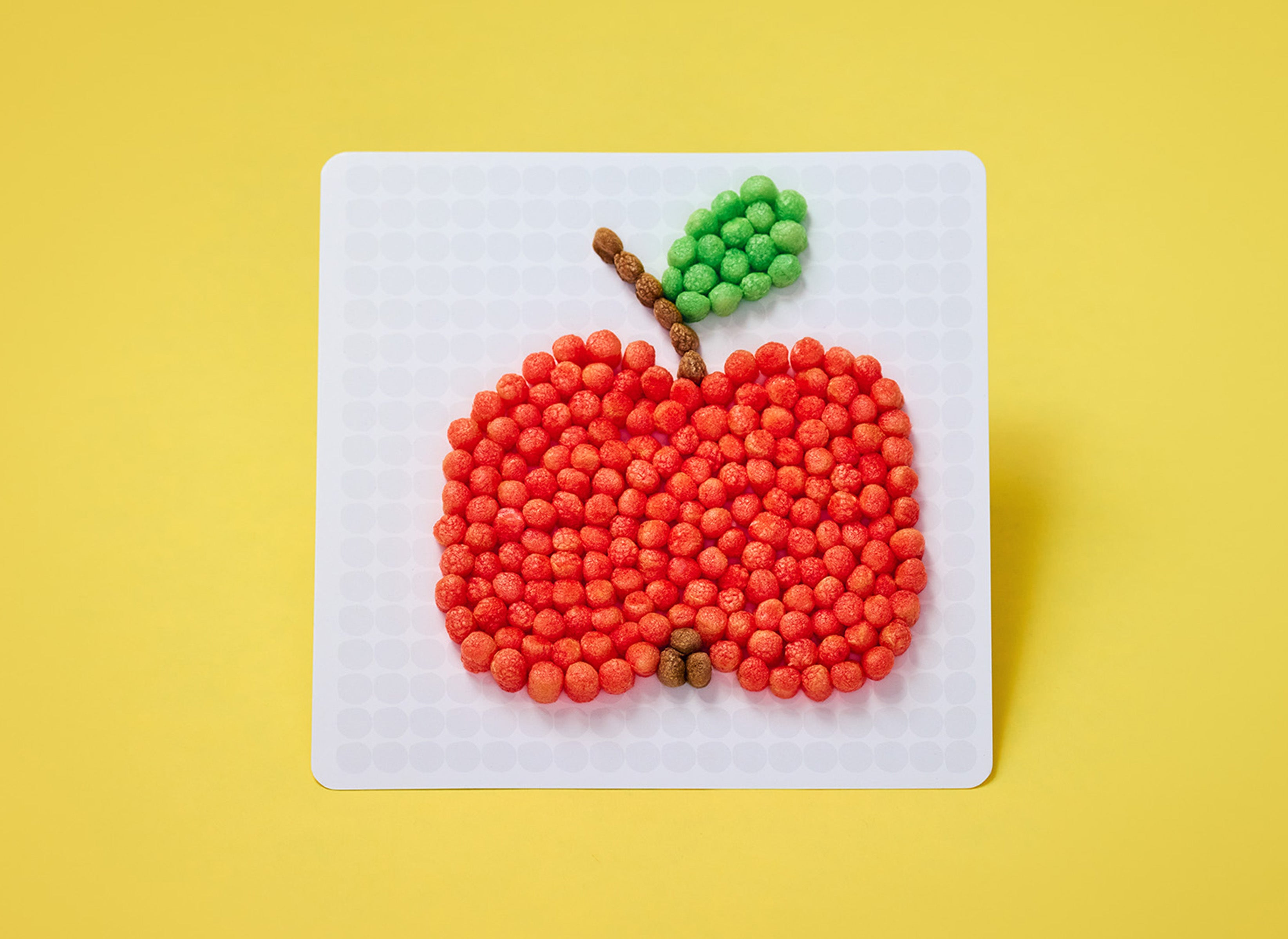 Ein Apfel aus roten, grünen und braunen Nubsis auf einer weißen Platte vor gelbem Hintergrund.