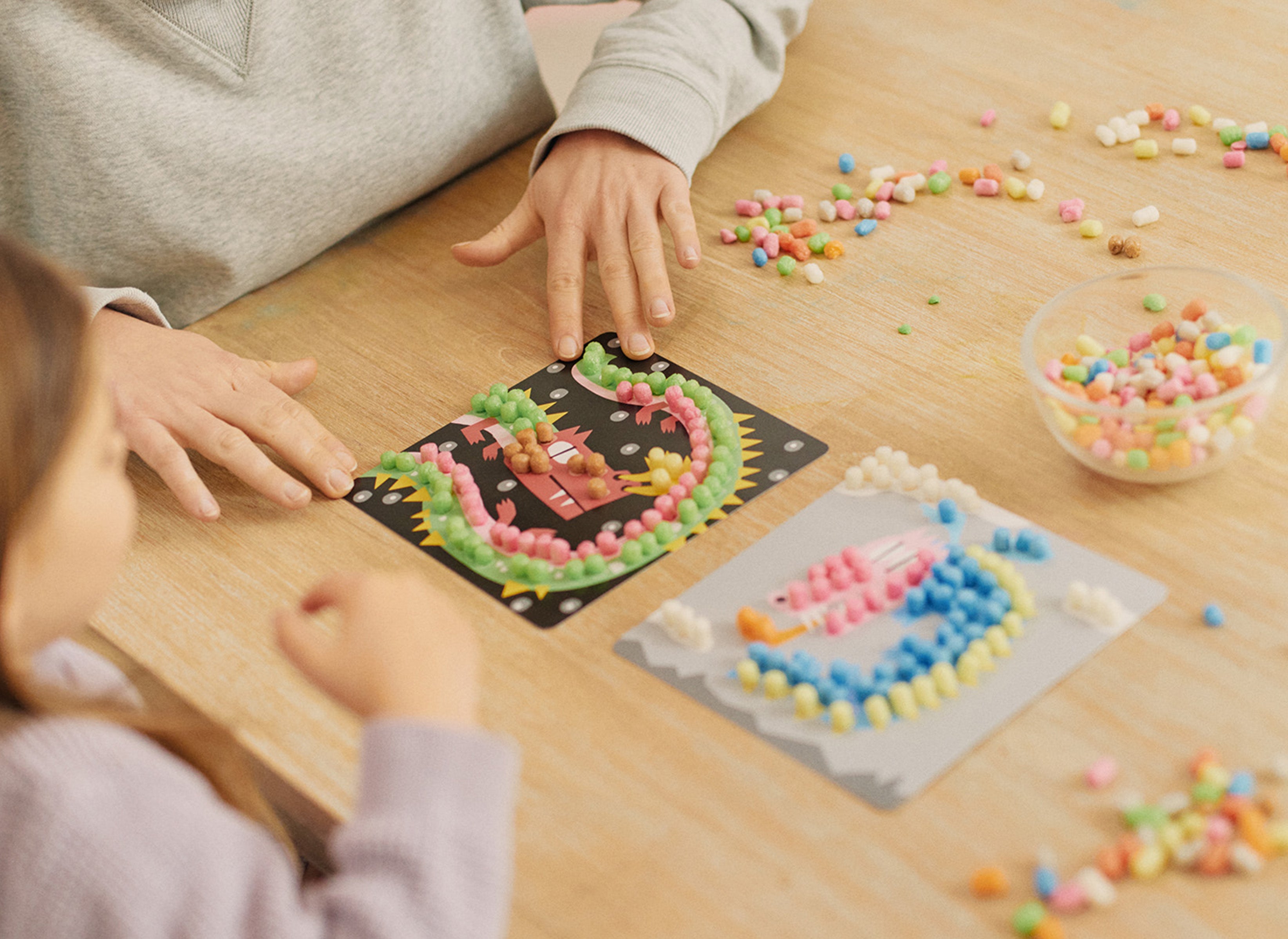 Person begleitet Kind beim Basteln mit bunten Nubsis auf Karten an einem Holztisch.