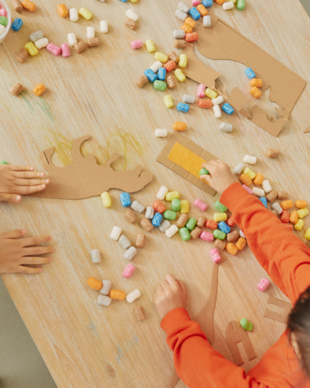 Kinder dekorieren Dinosaurierfiguren aus Karton mit bunten Nubsis auf einem Holztisch.