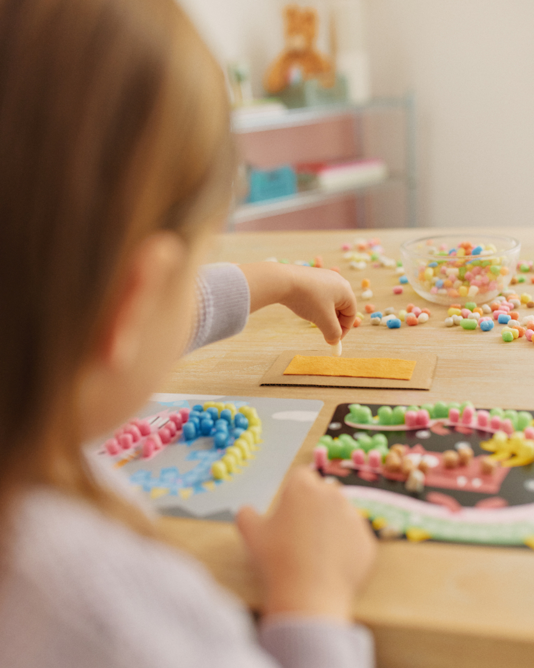 Ein Kind bastelt am Tisch mit bunten Nubsis, es klebt sie auf Papierkarten. Im Hintergrund stehen ein Glas und Spielzeug.