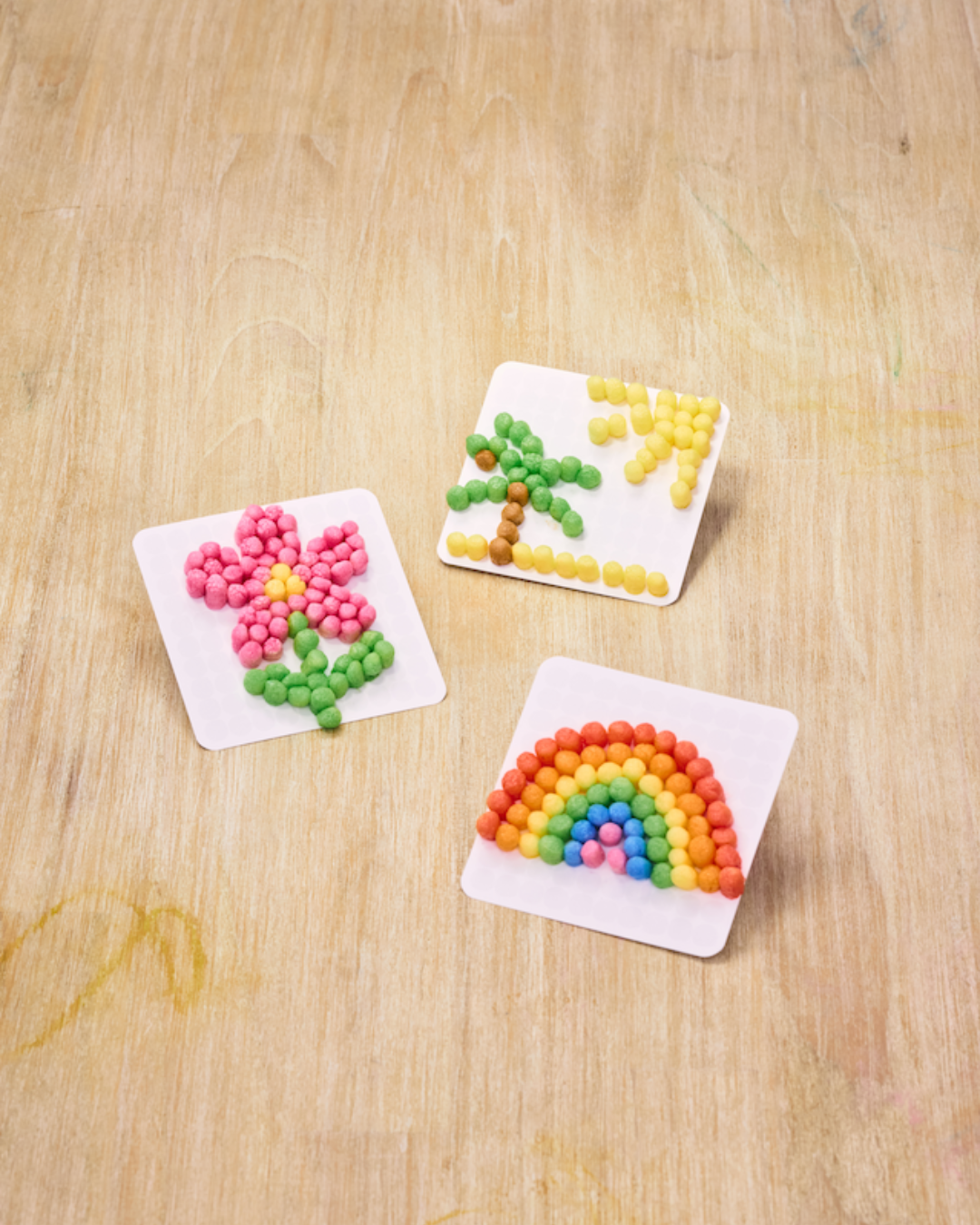 Drei Karten mit bunten Bildern aus Nubsis: eine Blume, eine Palme mit Sonne und ein Regenbogen, auf Holztisch.