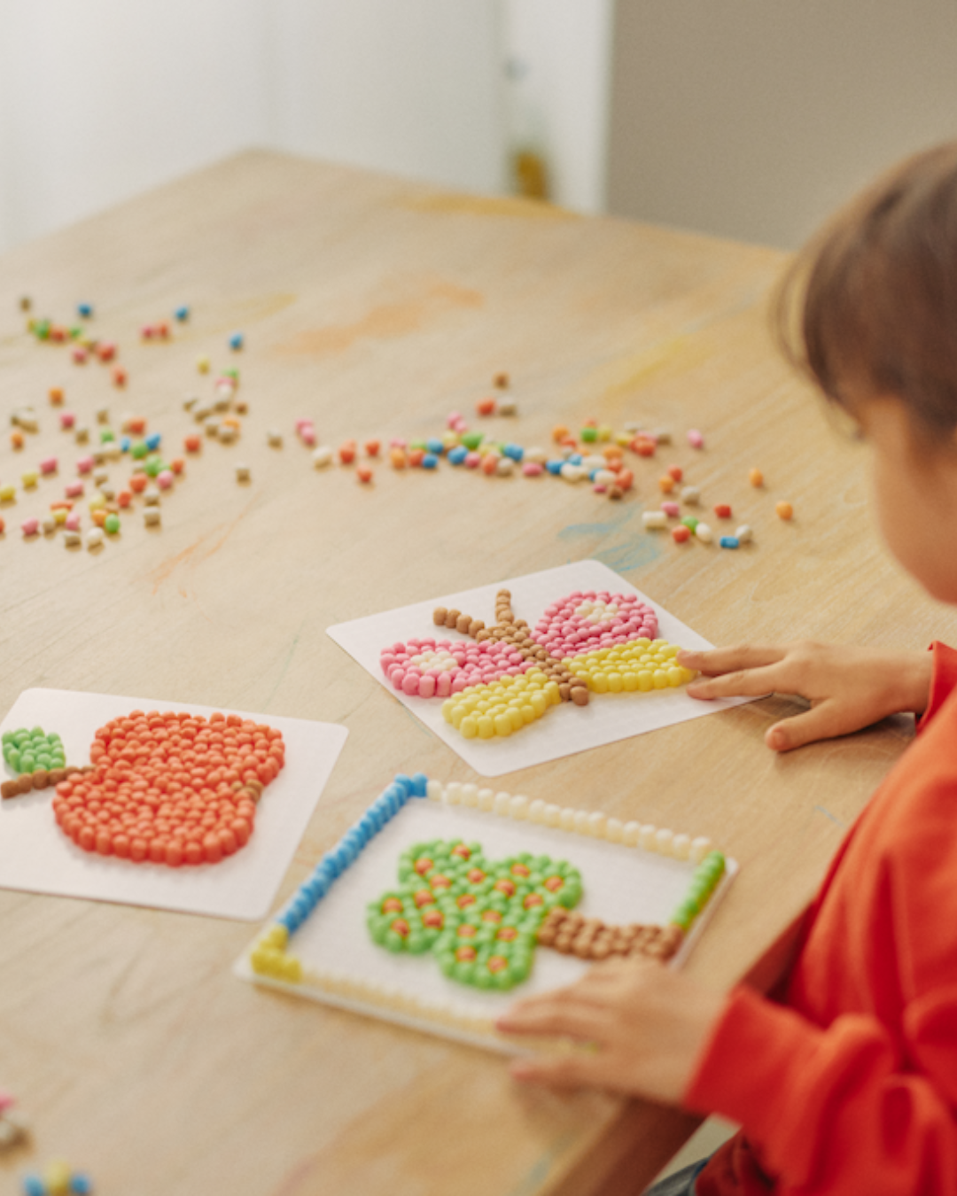 Ein Kind sitzt am Tisch und bastelt mit bunten Nubsis ein Bild von einem Apfel, Schmetterling und Baum auf Blattpapier.
