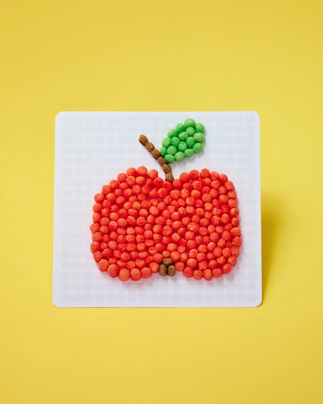 Ein Apfel aus roten, braunen und grünen Nubsis auf einer weißen Unterlage vor gelbem Hintergrund.