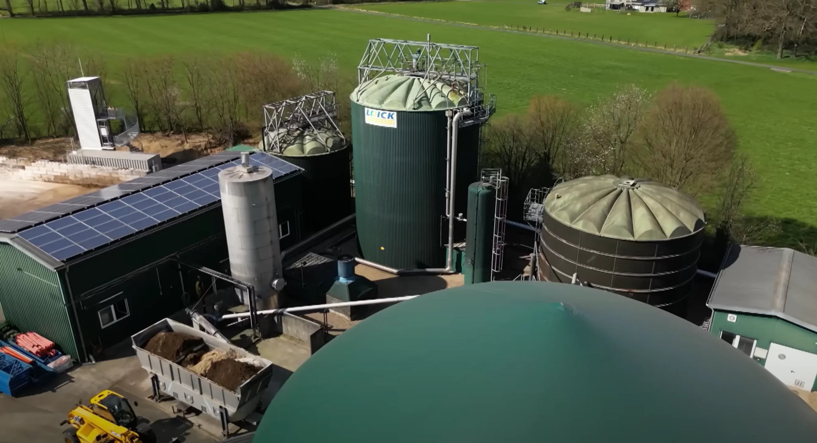 Luftbild einer Biogasanlage mit grünen Tanks, Solarpanels und umliegenden Feldern.