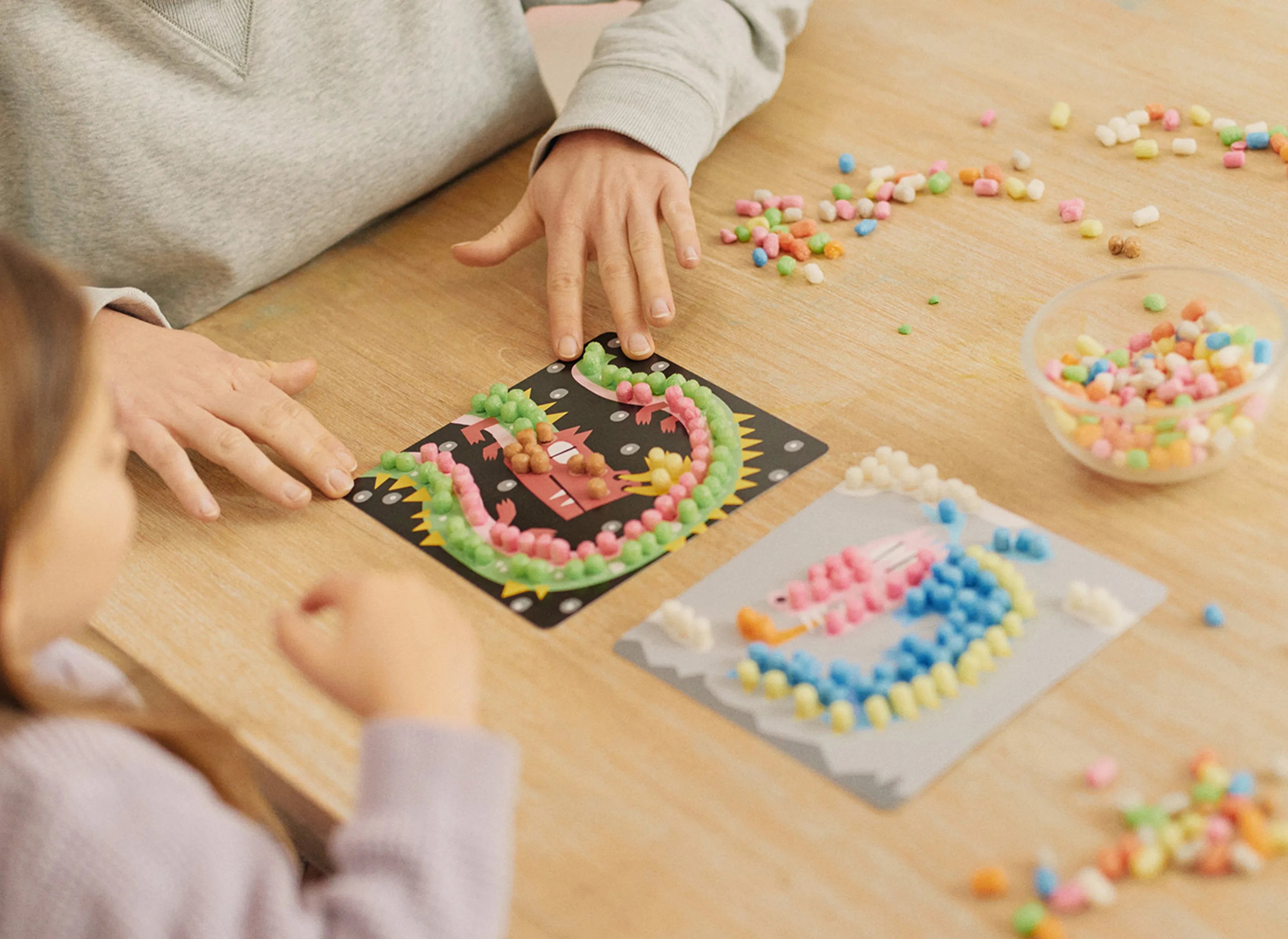 Personen basteln an einem Tisch mit bunten Playmais-Teilen auf Motiv-Vorlagen