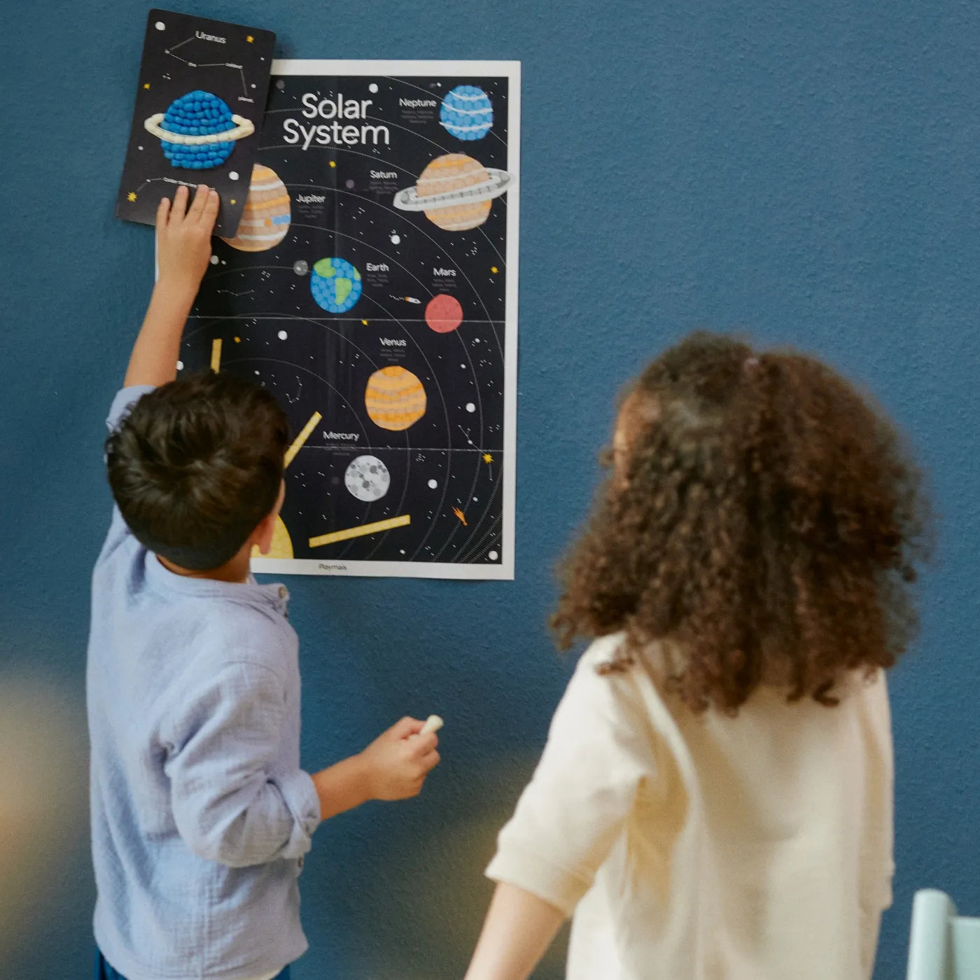 Zwei Kinder stehen vor dem Solar System Poster von Playmais. Ein Kind hält die Playmais Karte von Uranus an das Poster. Die Karte ist mit Playmais Mosaic beklebt.